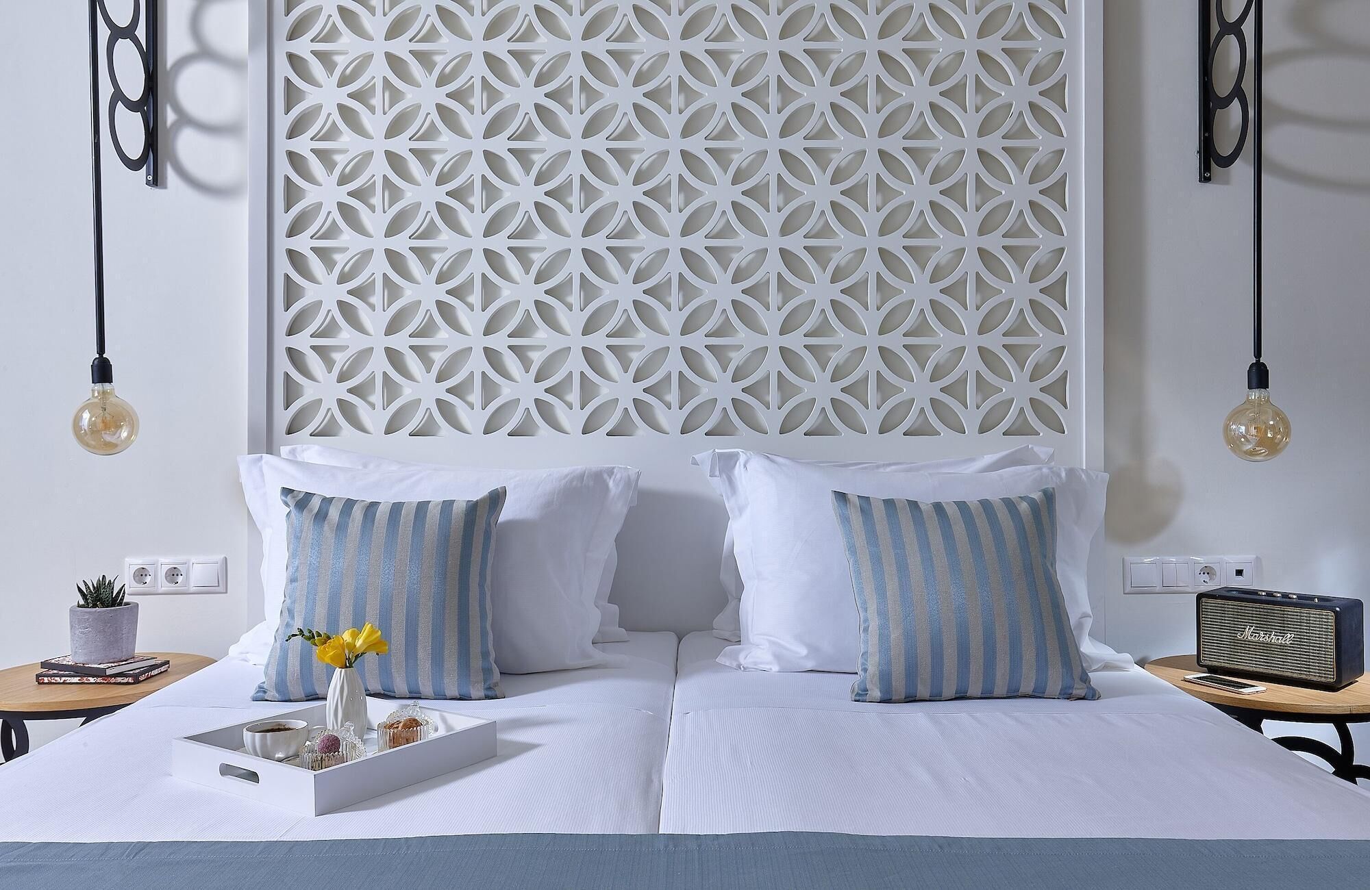 Una cama bien ordenada con ropa de cama blanca, almohadas a rayas, una bandeja decorativa con flores y una radio antigua sobre una mesita de noche.
