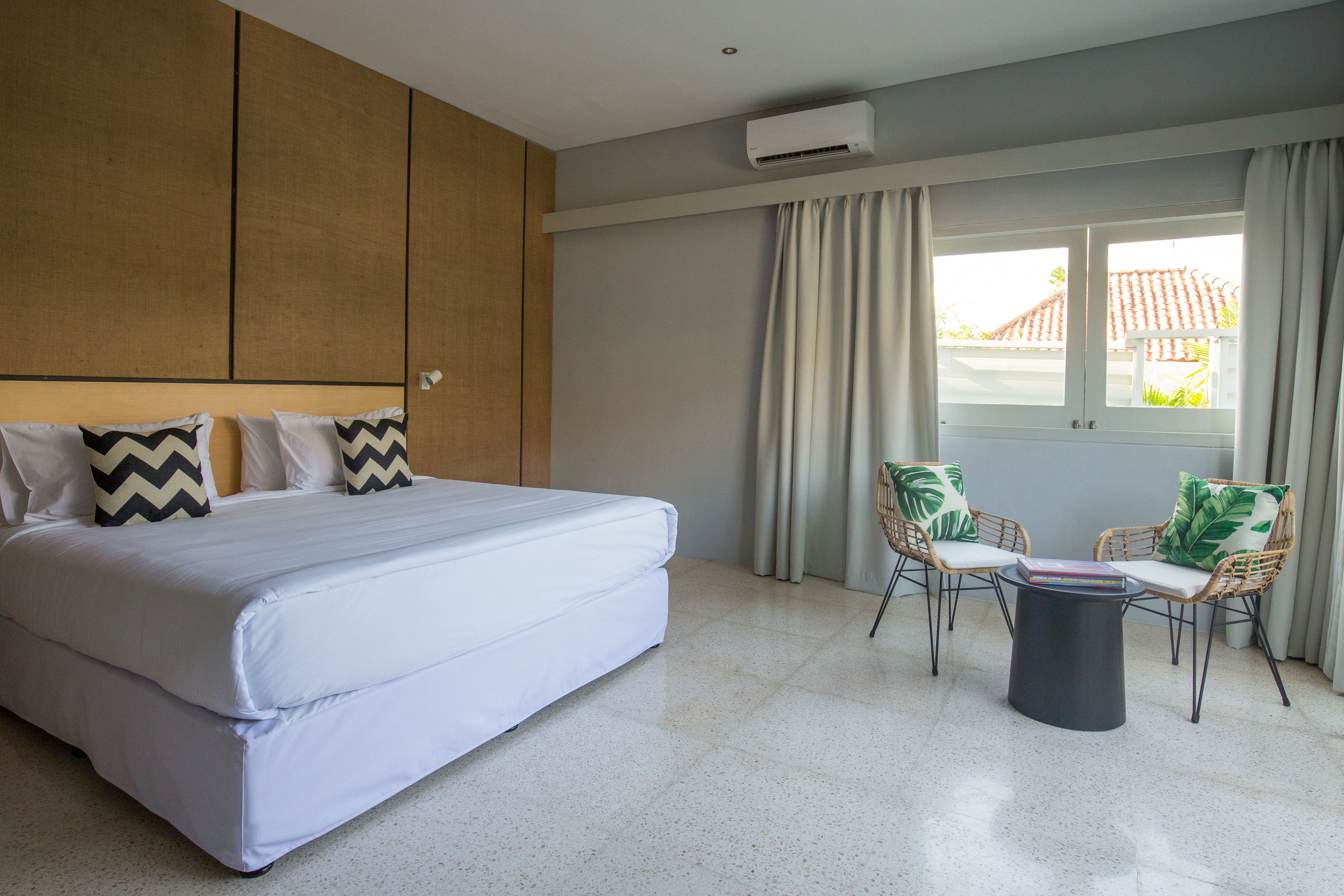 A modern hotel room with a large bed, two wicker chairs, a small table, and a window with curtains.