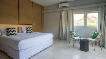 A modern hotel room with a large bed, two wicker chairs, a small table, and a window with curtains.