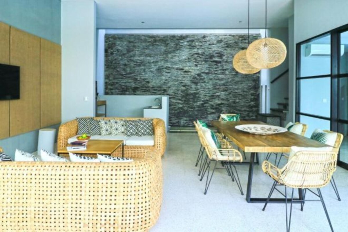A modern living room with a sofa, dining table, and chairs. There is a large window and a wall-mounted TV.