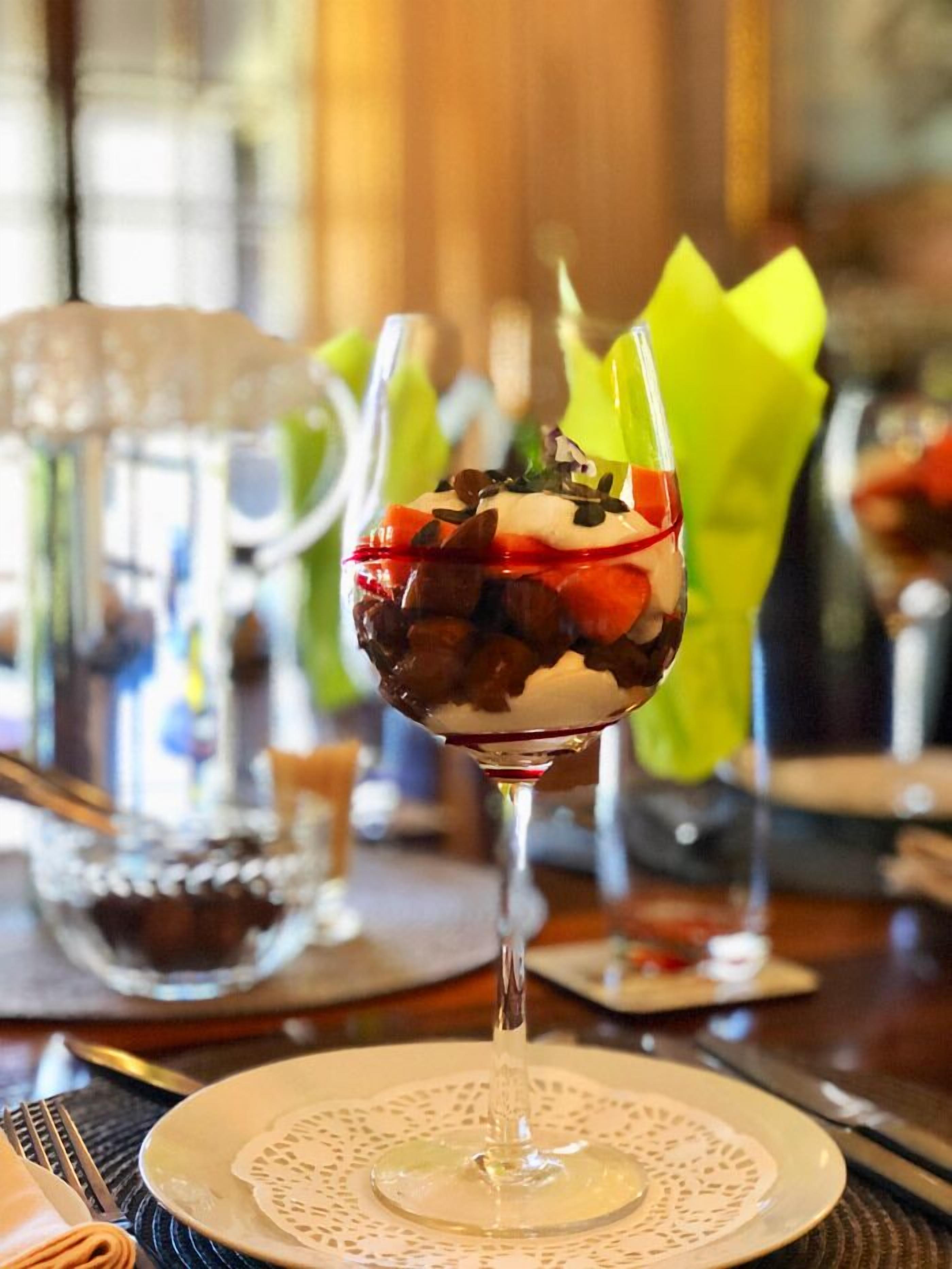 A dessert in a wine glass on a table with a doily, a green napkin, and a glass pitcher in the background.