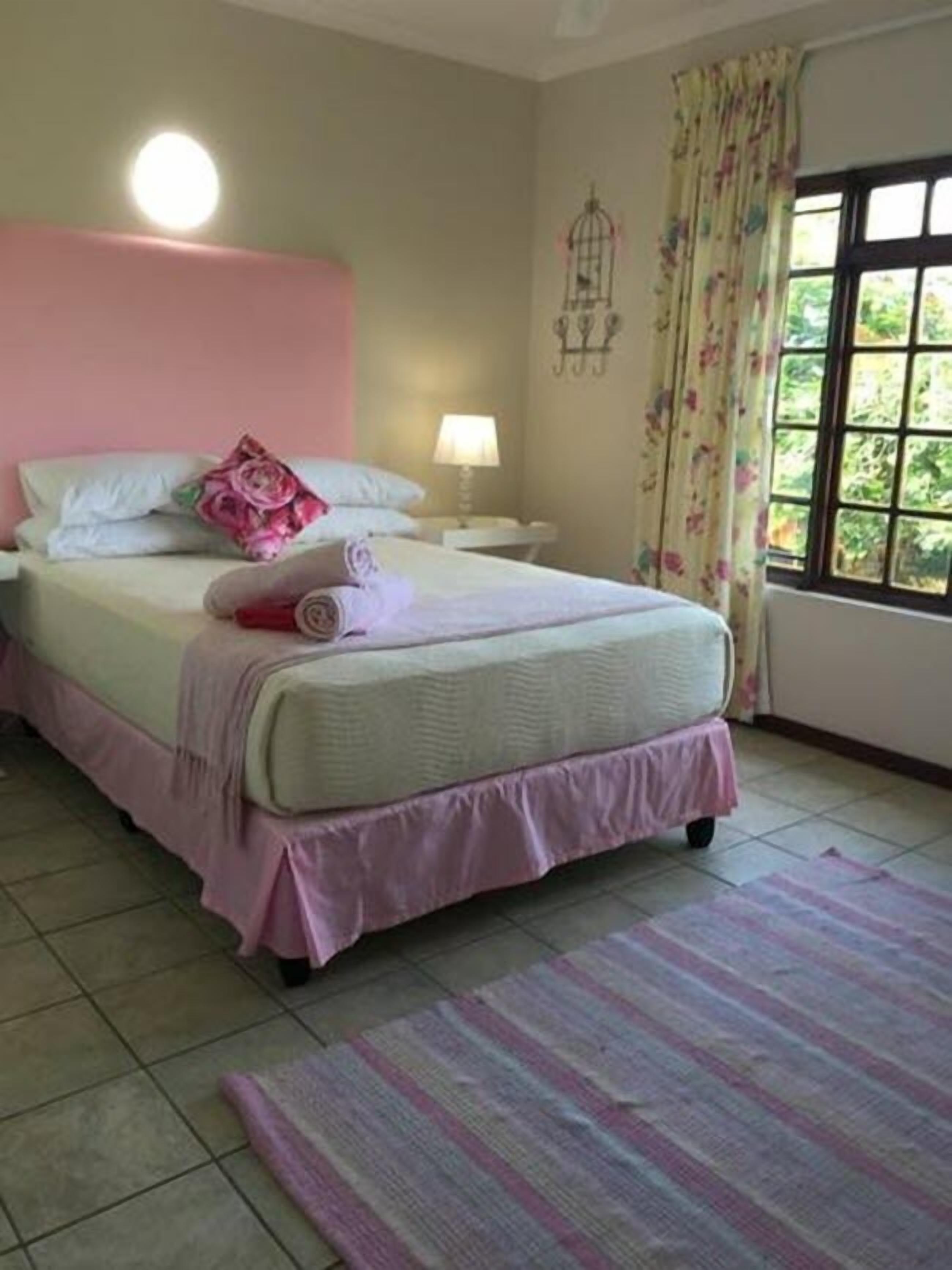 Una cama bien hecha con ropa de cama rosa, una lámpara de noche y una cortina con motivos florales.