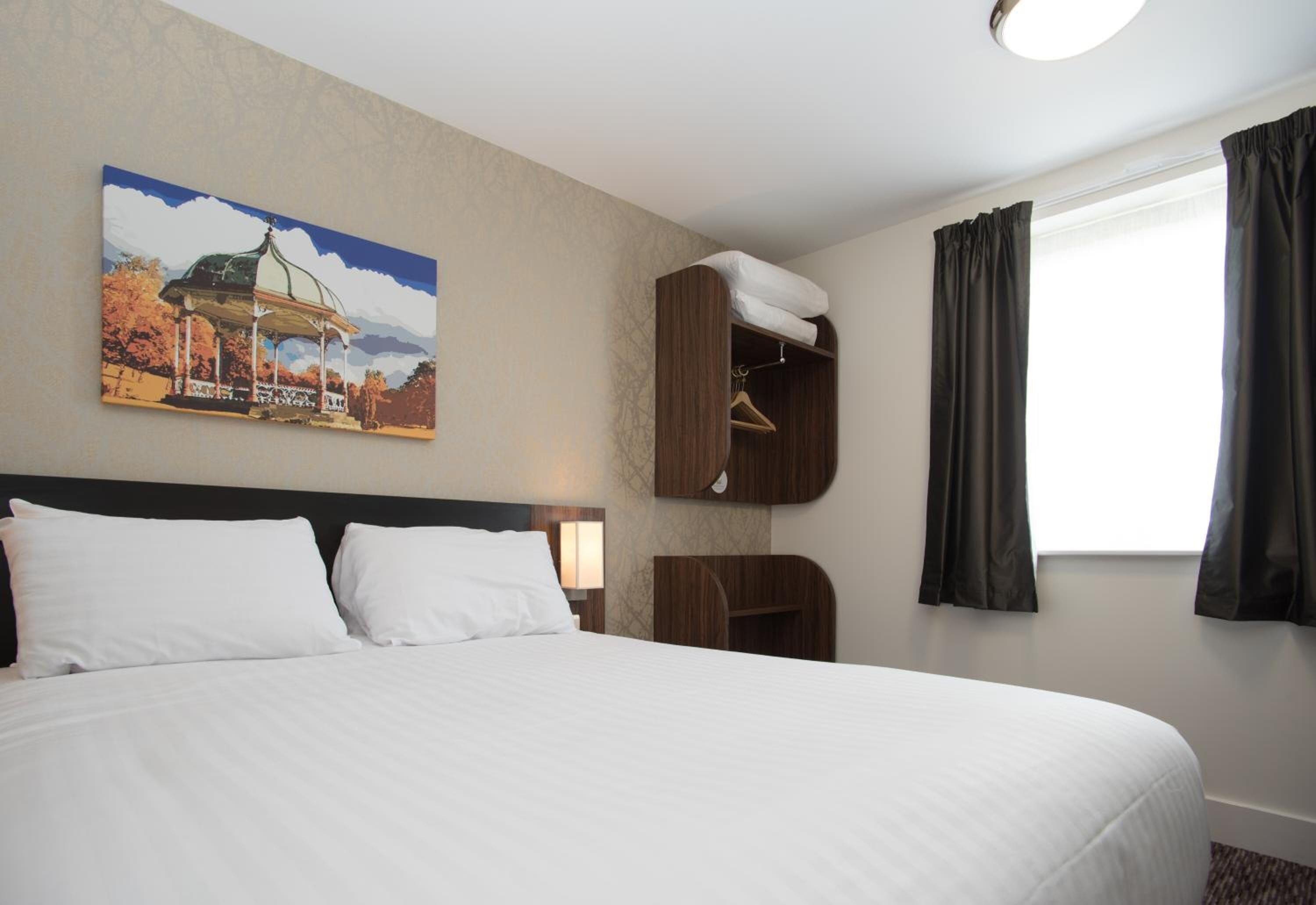 A hotel room with a large bed, a wooden wardrobe, a painting of a gazebo, and a window with curtains.