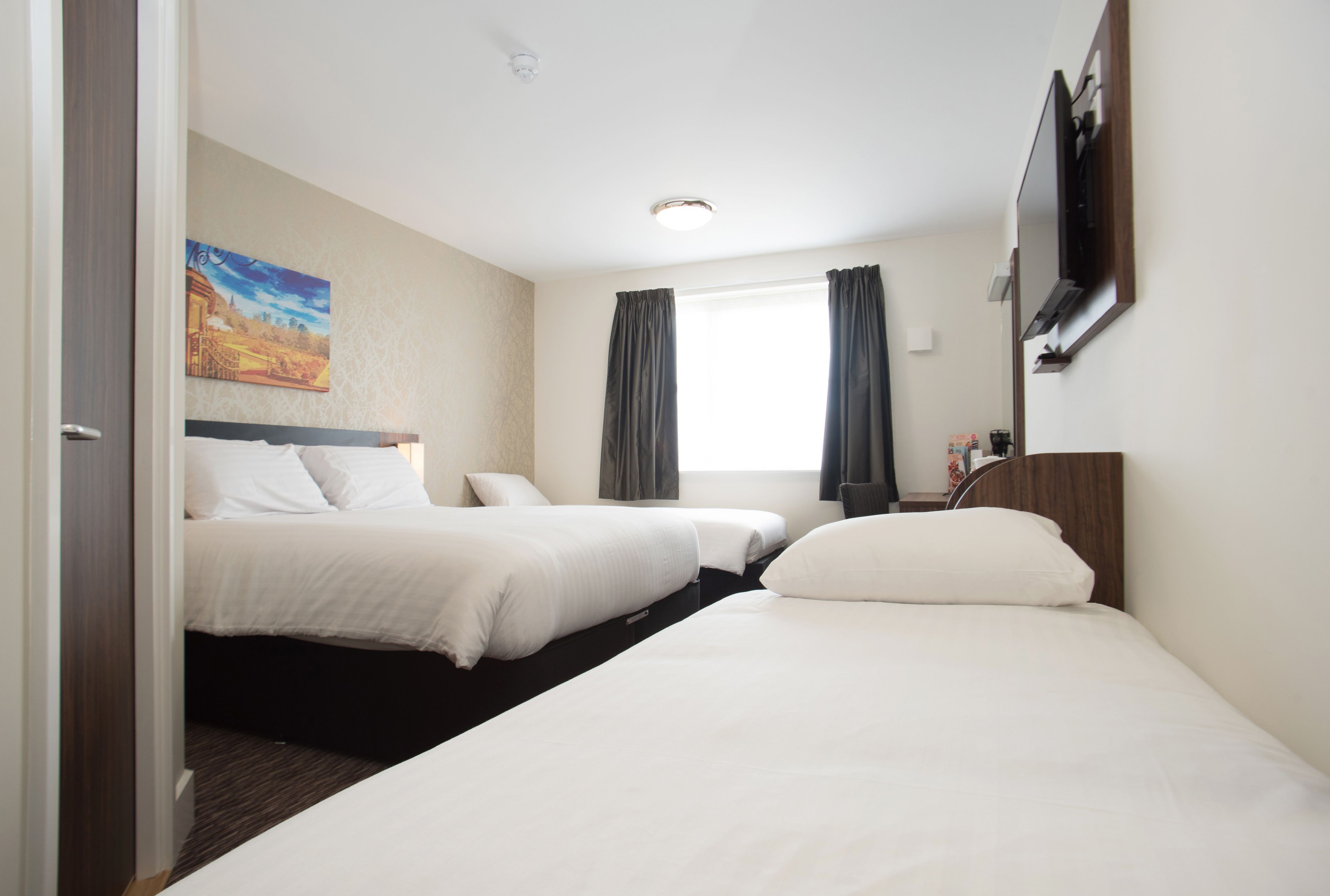 Una habitación de hotel con dos camas, un cuadro en la pared, una ventana con cortinas y un televisor de pantalla plana montado en la pared.