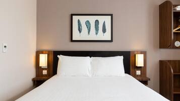 A hotel room with a large bed, two bedside lamps, a framed artwork of feathers, and a wooden nightstand with a shelf.