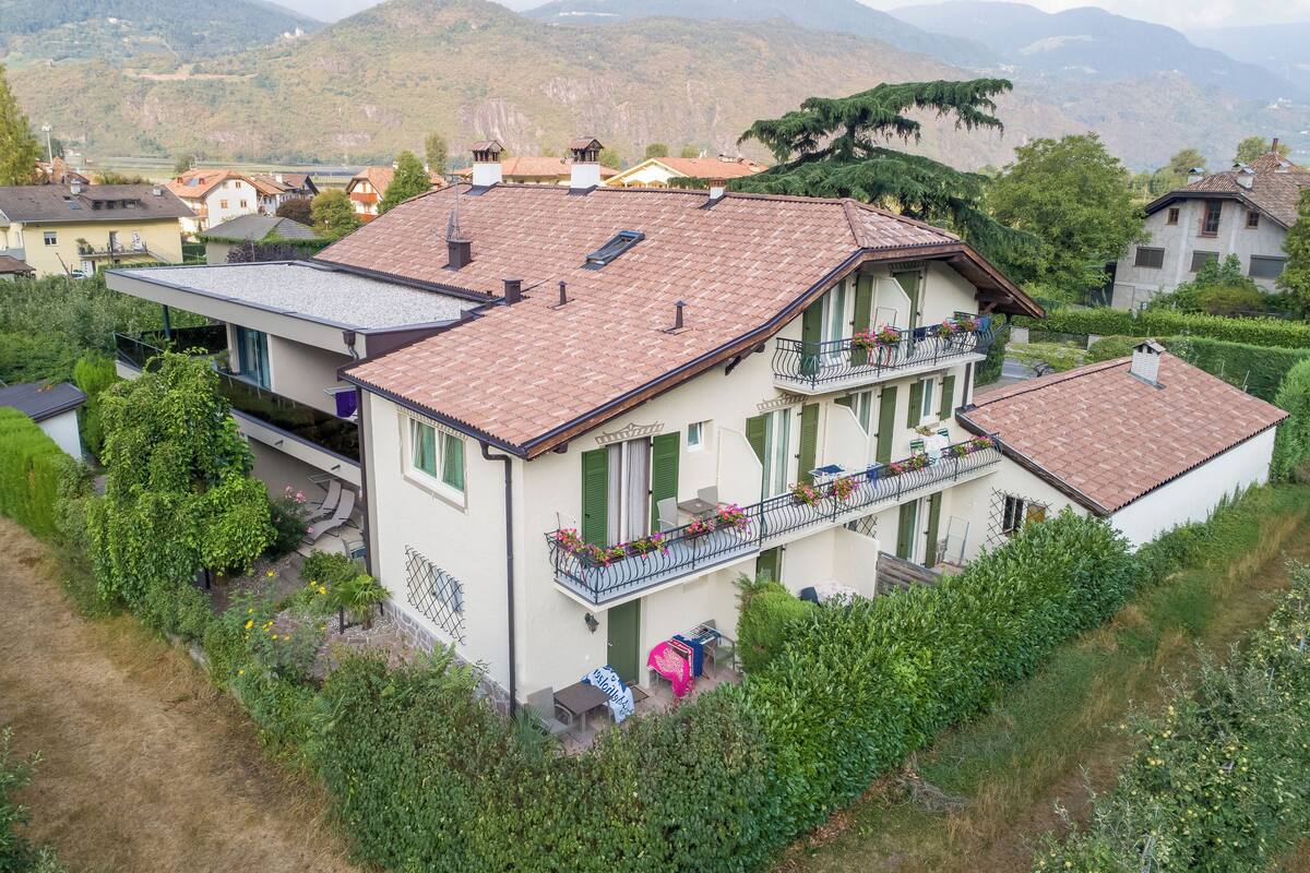 A two-story house with a balcony, surrounded by greenery and mountains in the background.