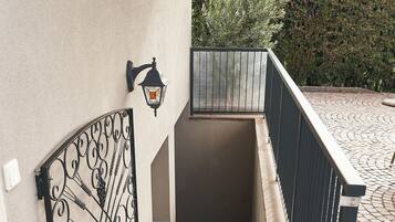 Un conjunto de escalones exteriores con barandilla de metal, una lámpara decorativa fijada a la pared y un felpudo metálico al final.