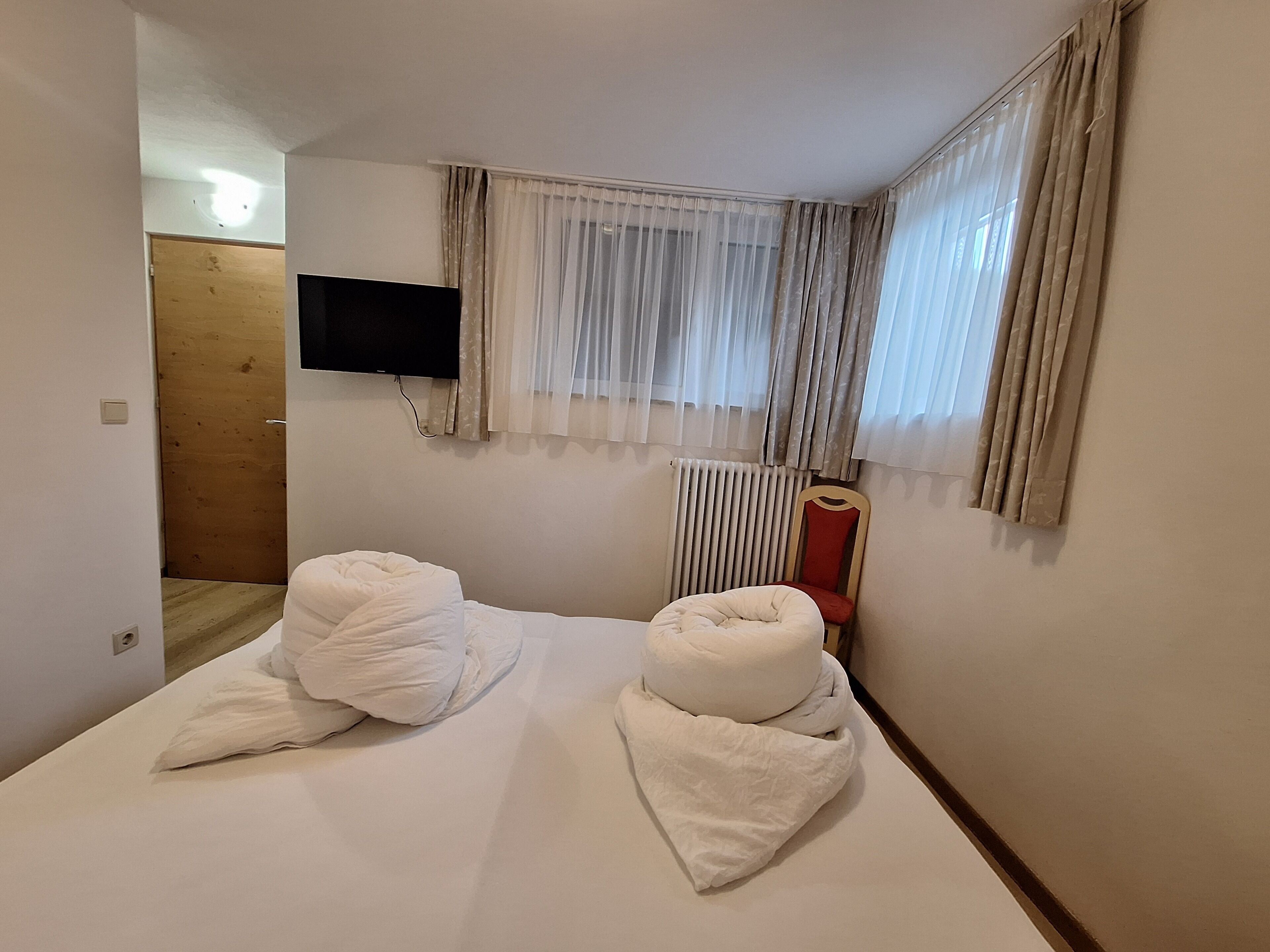 A hotel room with a bed, a television mounted on the wall, a radiator, and a window with curtains.
