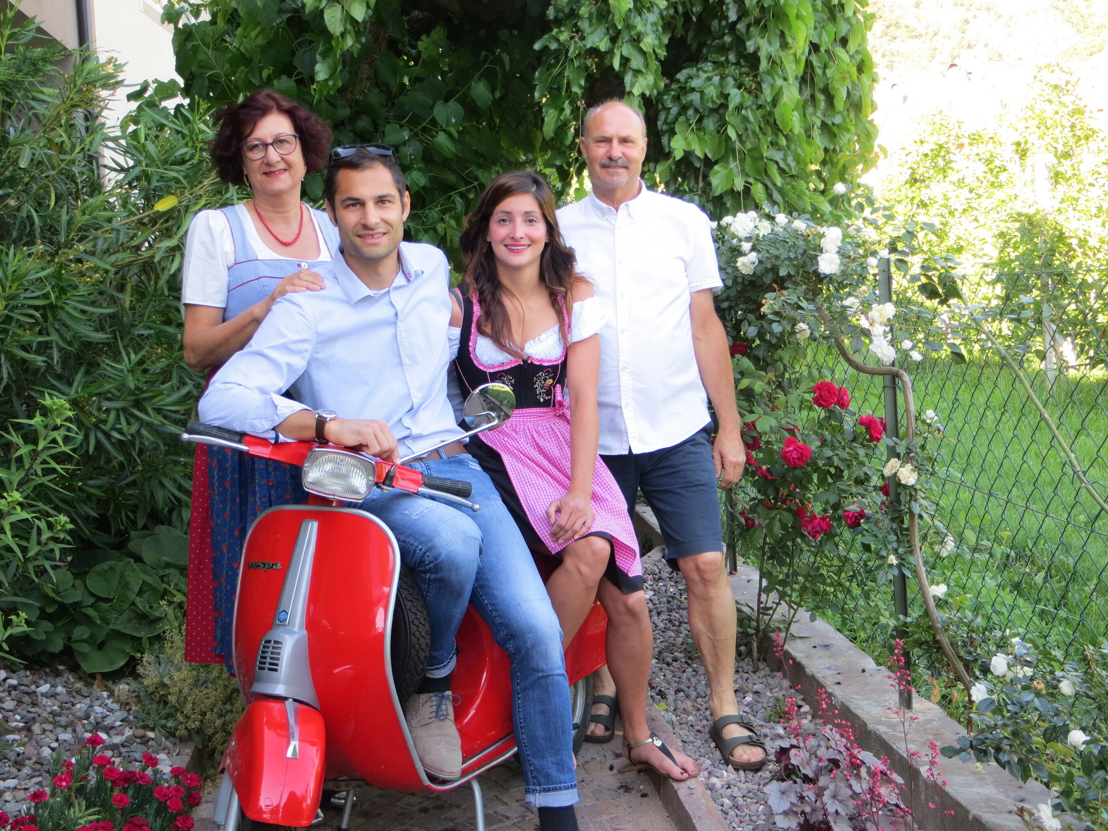 A family on a red scooter in a garden with a fence and flowers.
