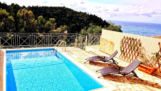 A rectangular pool with clear blue water, surrounded by a stone deck and a metal railing. There are two lounge chairs and a potted plant on the deck. - Agios Nikitas View (Lefkada)