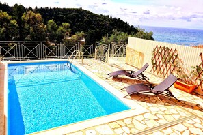 A rectangular pool with clear blue water, surrounded by a stone deck and a metal railing. There are two lounge chairs and a potted plant on the deck. - Agios Nikitas View (Lefkada)