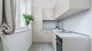 A modern kitchen with light-colored cabinets, a herringbone-patterned backsplash, and a window with a curtain. - Elite Apartments King Size Old Town (Gdansk)