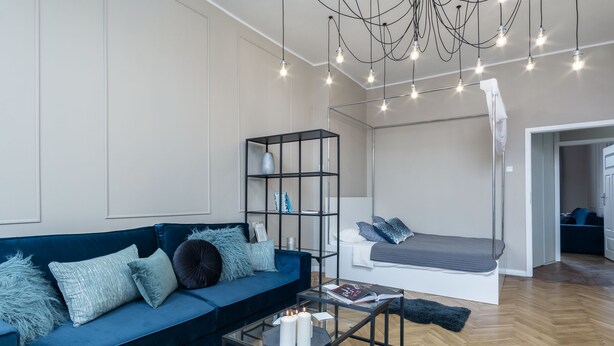 A modern living room with a blue sofa, a glass coffee table, a white bed, and a black shelving unit. - Elite Apartments King Size Old Town (Gdansk)