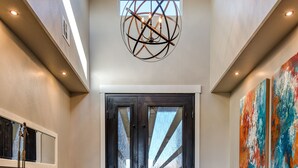 En rymlig entréhall med trägolv, en stor matta, en mörk träbänk, en modern kristallkrona och ett par dubbeldörrar med en geometrisk glasdesign.