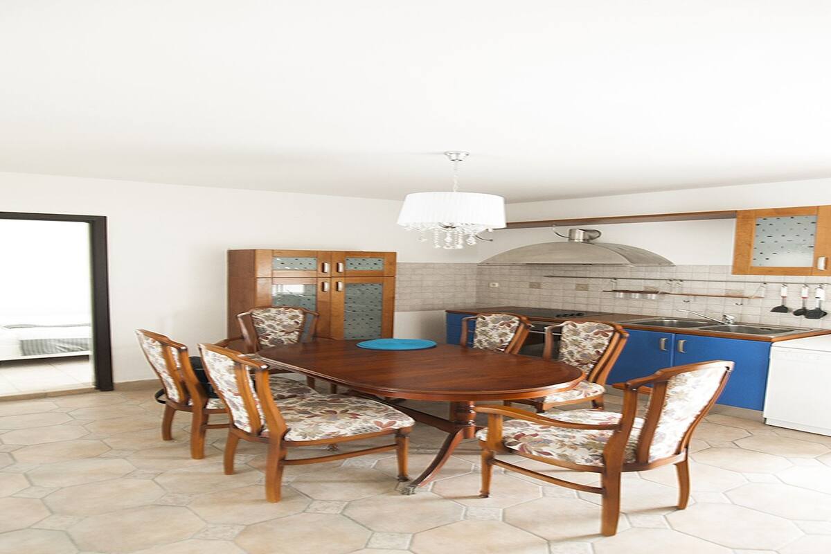 Un comedor con una mesa y sillas de madera, una lámpara de araña, y una cocina con armarios azules y una encimera blanca.