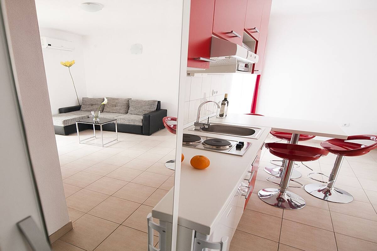 Una cocina moderna con encimera blanca, taburetes rojos y un sofá gris en la sala de estar.
