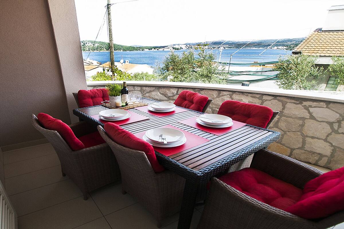Un balcón con una mesa para cuatro personas, sillas de mimbre con cojines rojos, y vistas al mar y edificios al fondo.