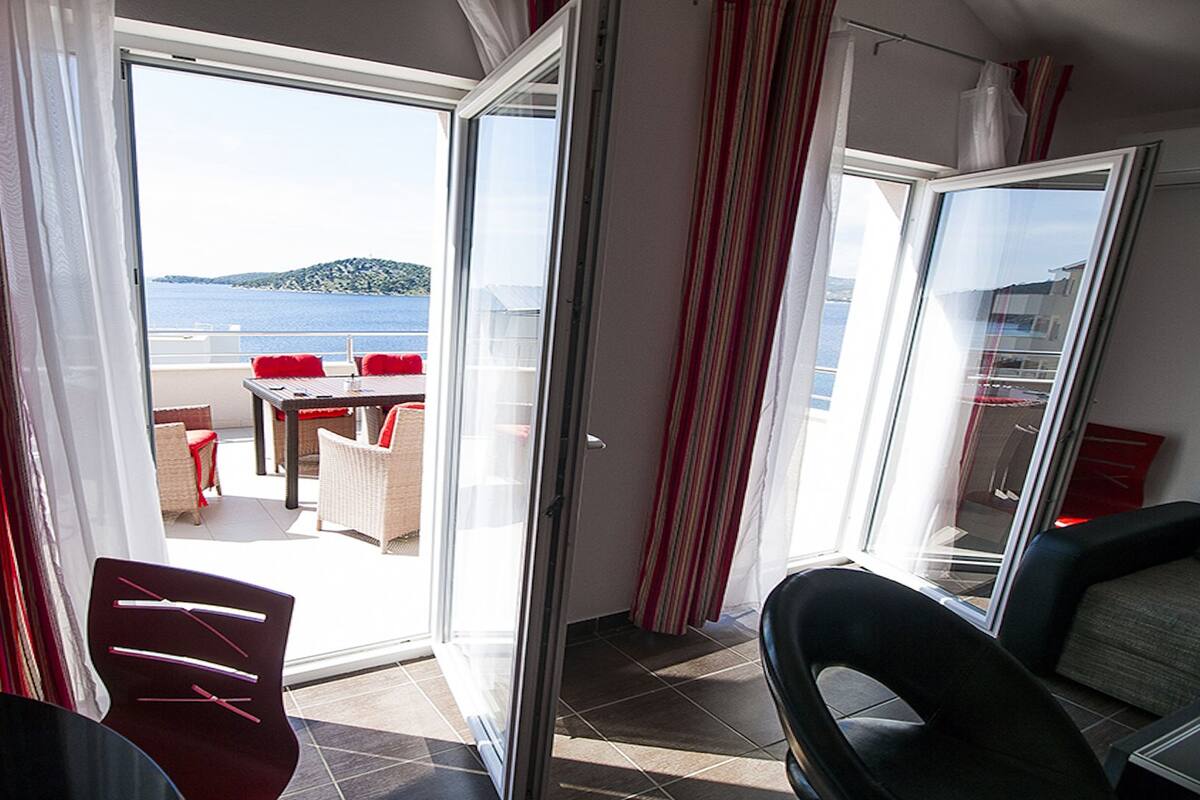 Habitación con puerta corrediza de vidrio que da a un espacio exterior con mesa y sillas, y vista al mar.
