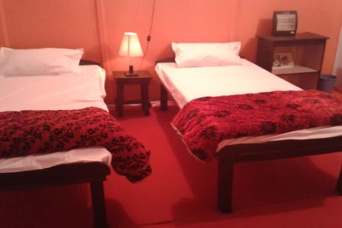 A hotel room with two beds, each with white linens and a red patterned blanket. There is a small bedside table with a lamp between the beds.