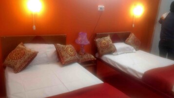 Two single beds with patterned pillows and red bedspreads in a room with orange walls and two wall-mounted lights.