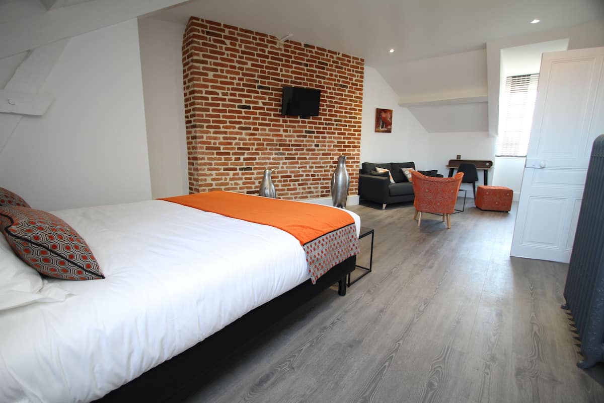 A modern hotel room with a brick accent wall, a large bed with an orange throw, a desk with a chair, and a television mounted on the wall.