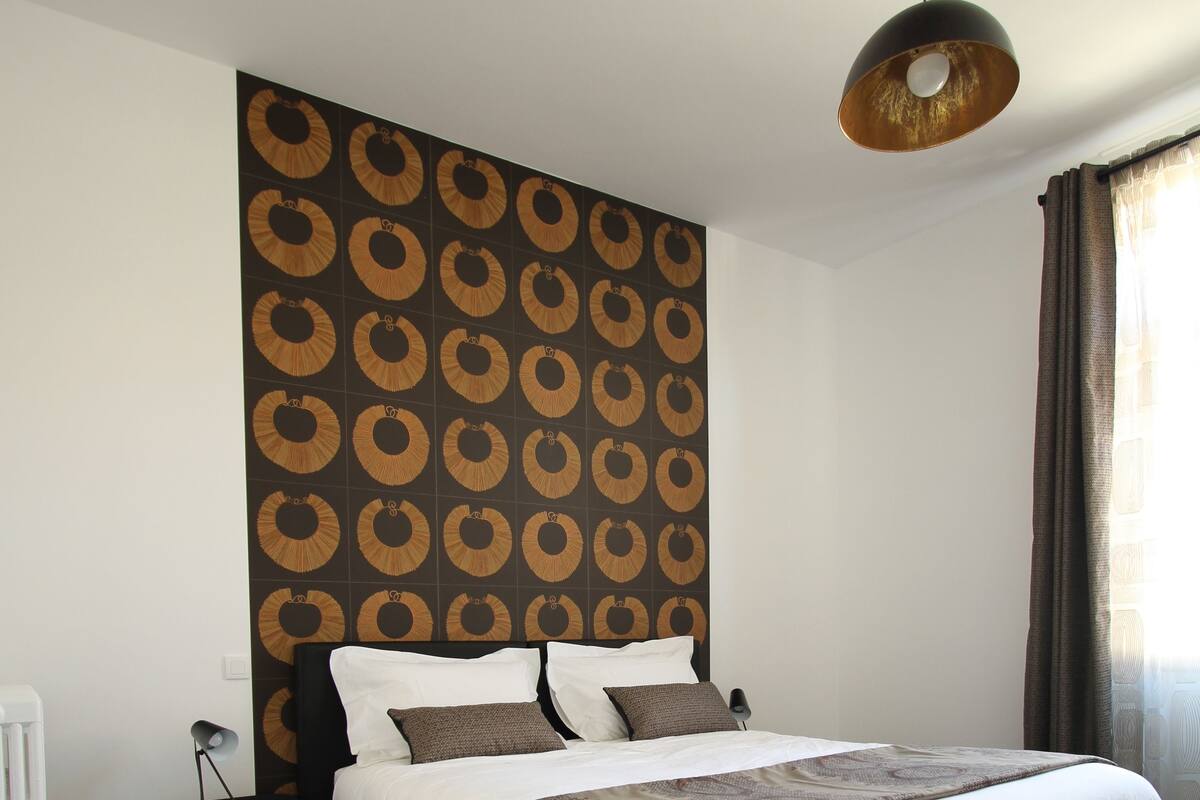 A neatly arranged bedroom with a large bed, a bedside table, a wall with a circular pattern, and a hanging light fixture.