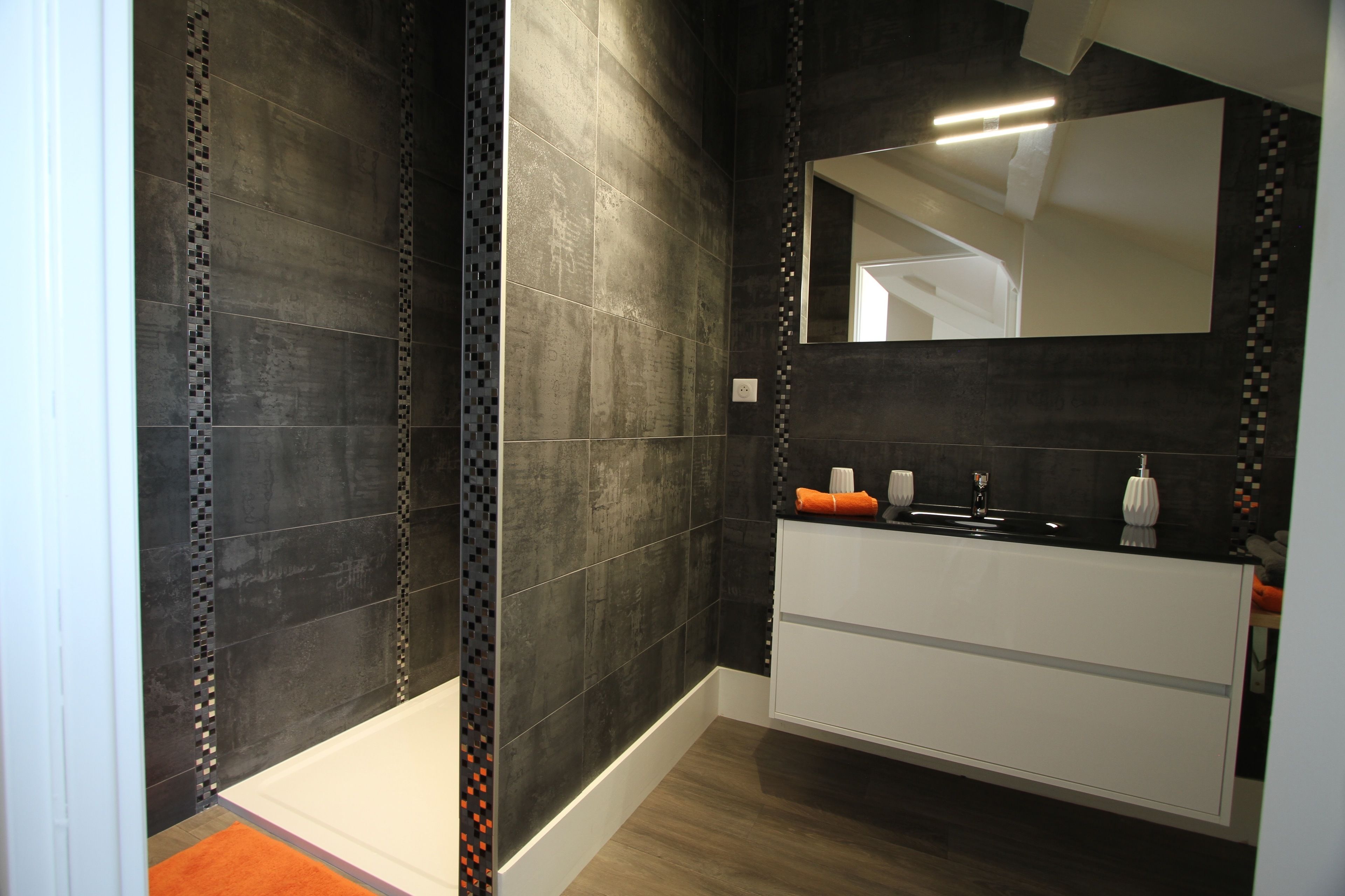 A modern bathroom with dark tiled walls, a white vanity, and a large mirror.