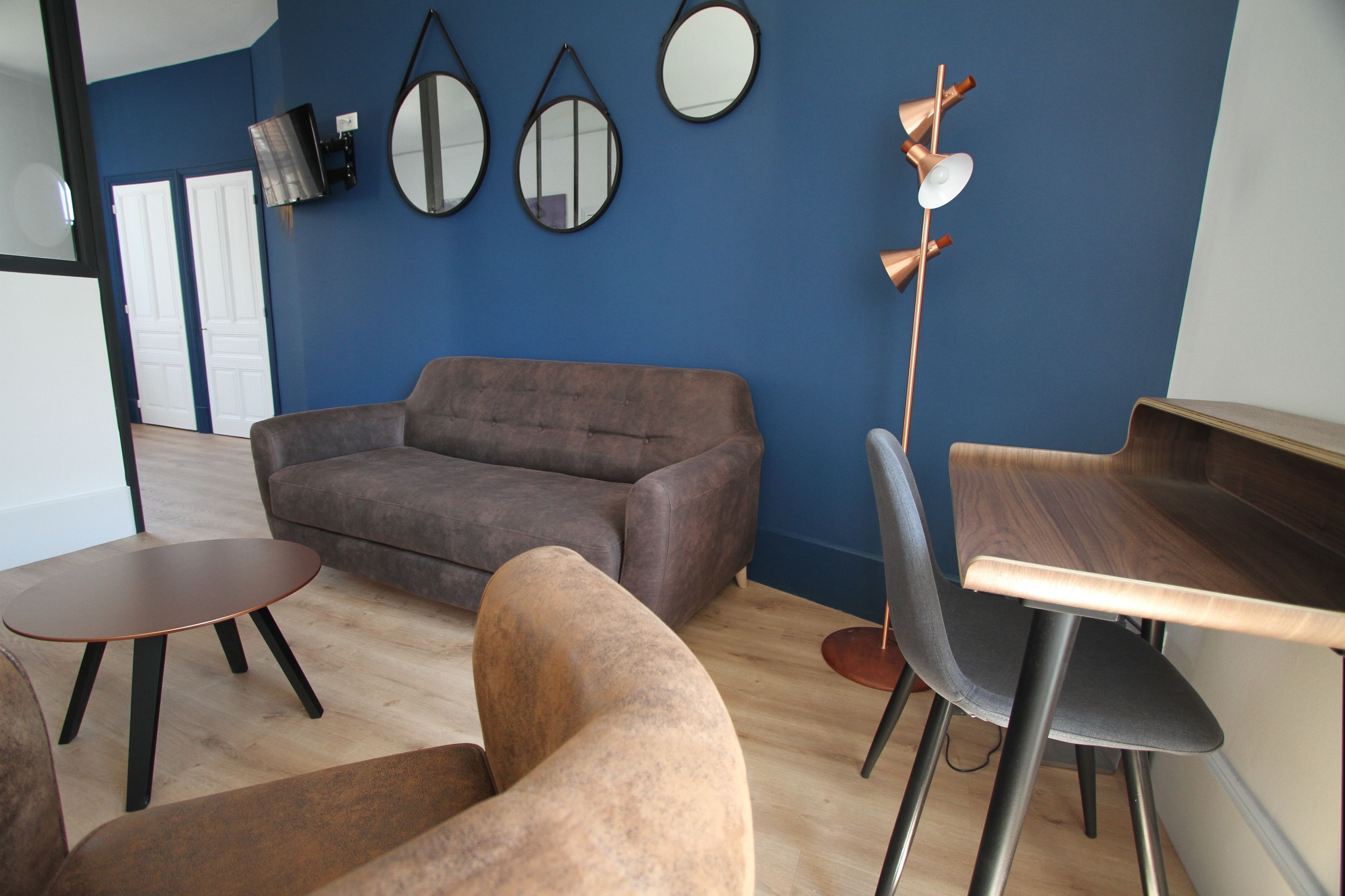 A modern living room with a brown sofa, a wooden coffee table, and a dining area with a wooden table and chairs. There are three wall-mounted mirrors and a tall floor lamp.
