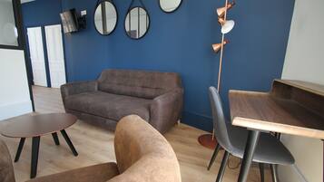A modern living room with a brown sofa, a wooden coffee table, and a dining area with a wooden table and chairs. There are three wall-mounted mirrors and a tall floor lamp.