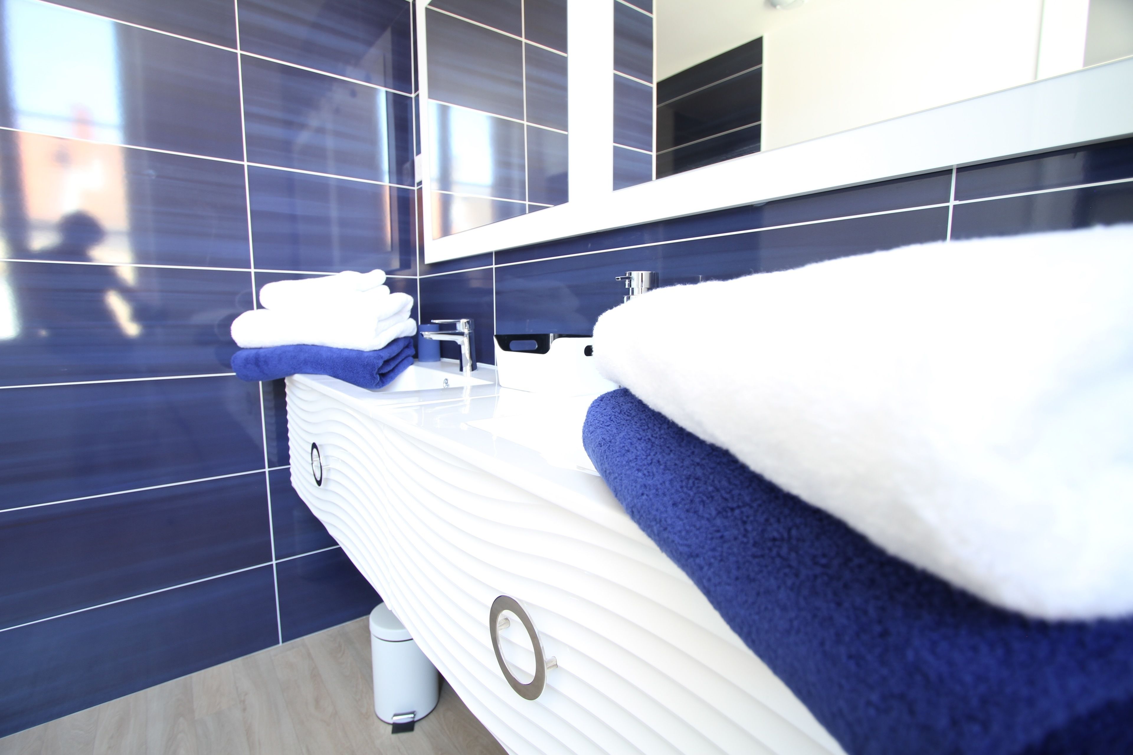 A modern bathroom with a white vanity, dark blue tiles, and a large mirror.