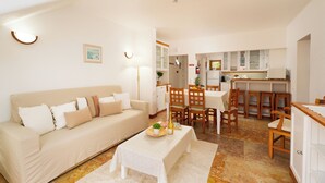 Una sala de estar con sofá, una mesa de comedor y sillas. Se observa una cocina al fondo con armarios y una encimera.