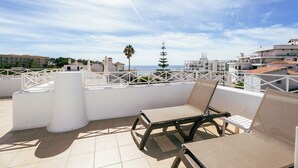Una terraza en la azotea con dos sillones beige, una barandilla blanca y vistas al mar y edificios.