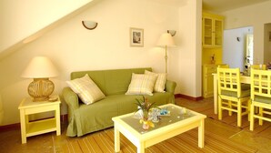 A cozy living room with a green sofa, two lamps, a coffee table, and a dining area with yellow chairs.