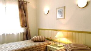 A hotel room with two beds, a bedside table with a lamp, a framed picture on the wall, and a window with curtains.