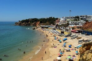 A beach with people swimming and sunbathing, boats parked on the shore, and buildings in the background.