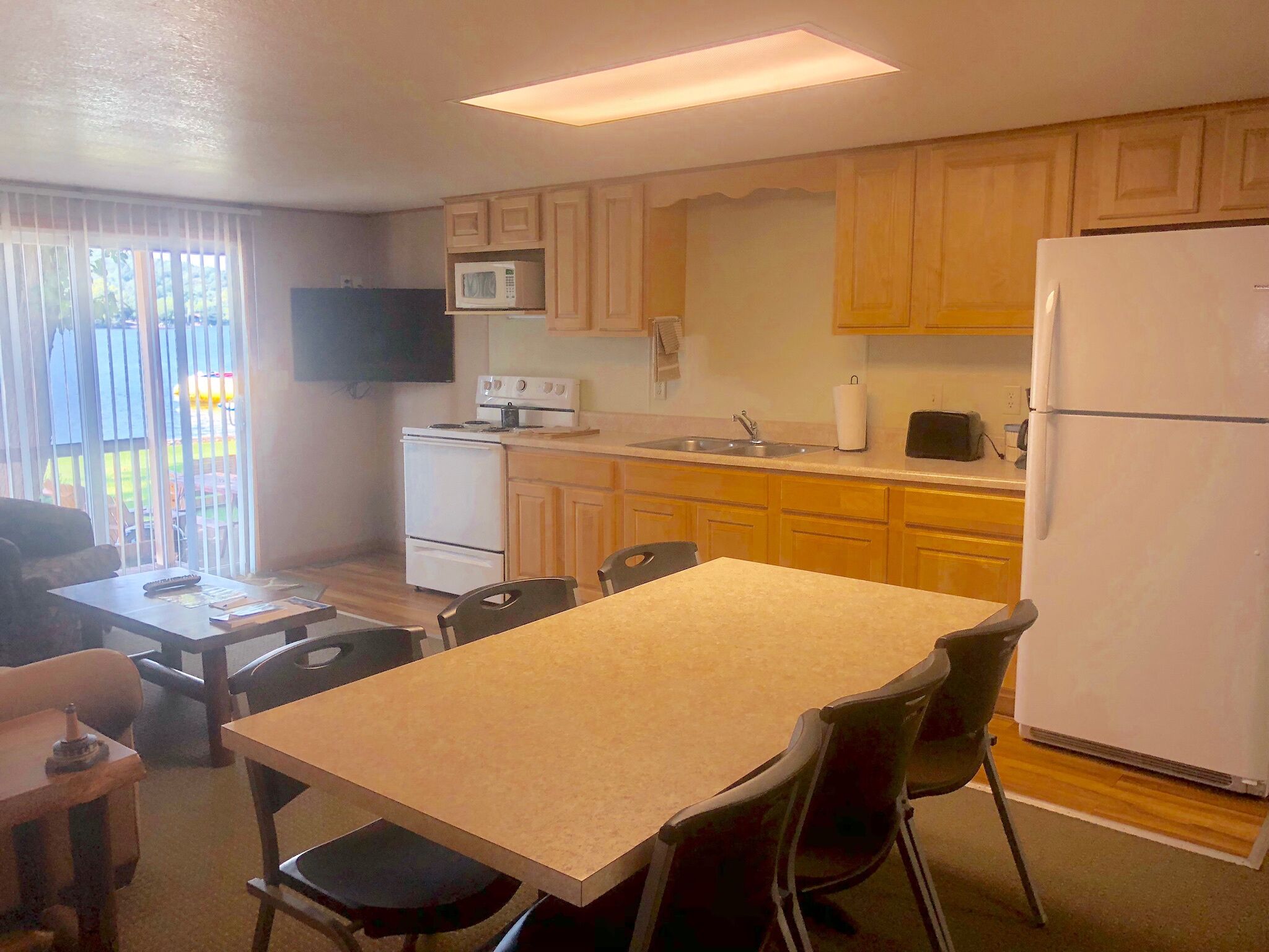 Una cocina compacta con armarios de madera, microondas, estufa, fregadero y refrigerador. Hay una mesa de comedor con sillas y un televisor montado en la pared.