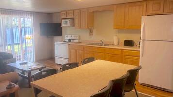 Una cocina compacta con armarios de madera, microondas, estufa, fregadero y refrigerador. Hay una mesa de comedor con sillas y un televisor montado en la pared.