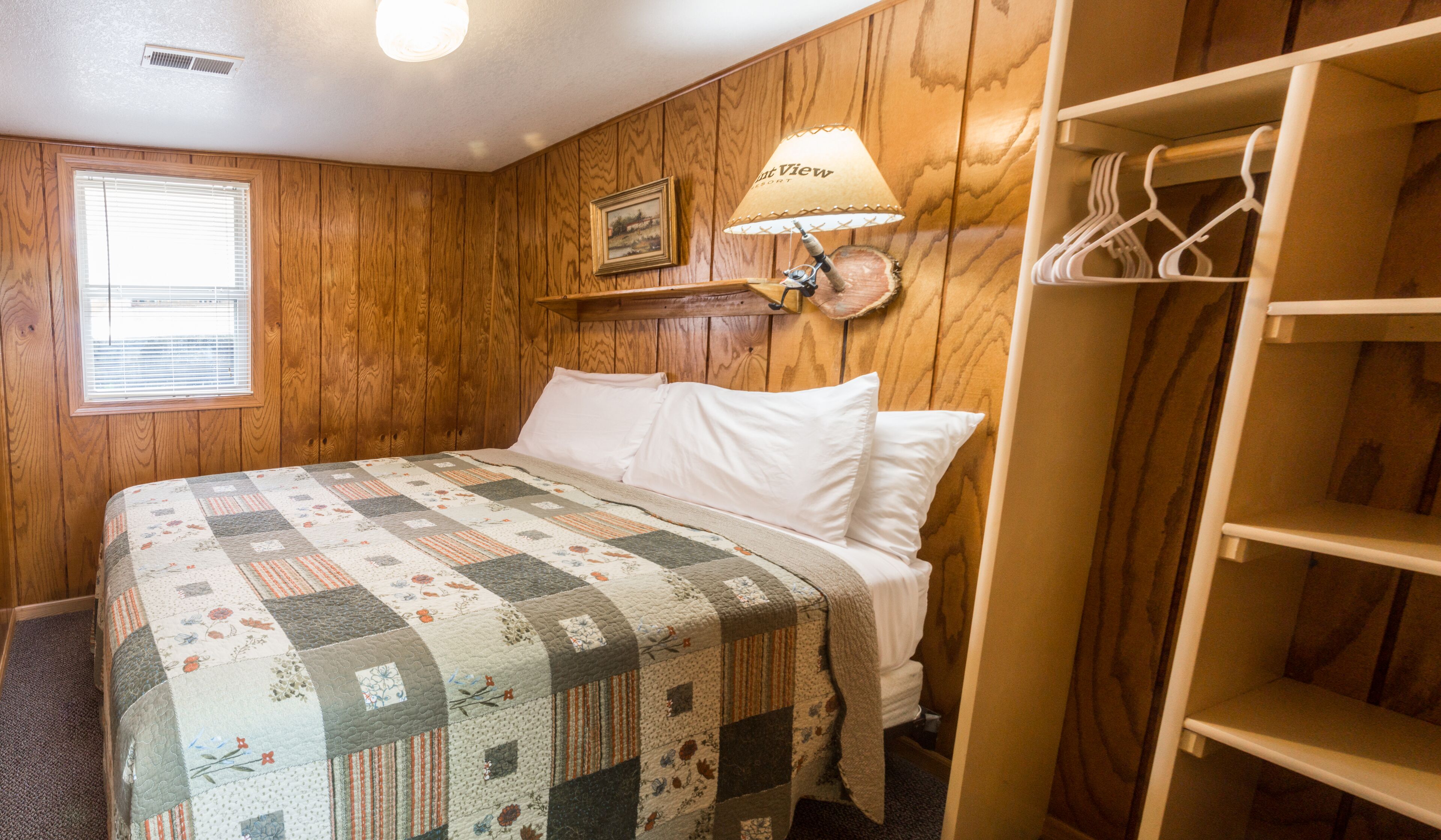 A bedroom with a wooden paneled wall, a quilted bedspread, a bedside lamp, a framed picture, and a wooden closet with hangers.
