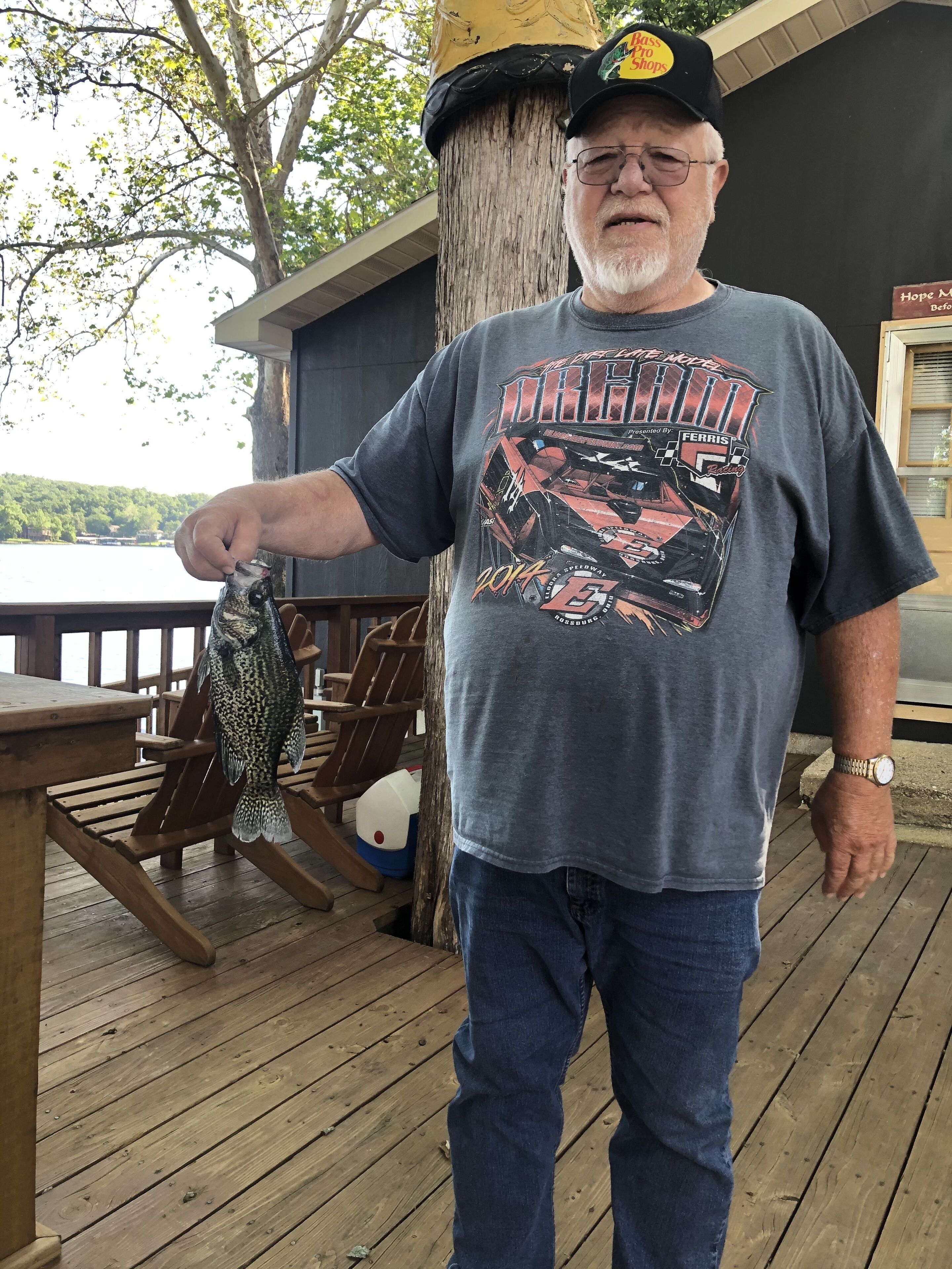 A man holding a fish on a wooden deck with a lake view in the background.