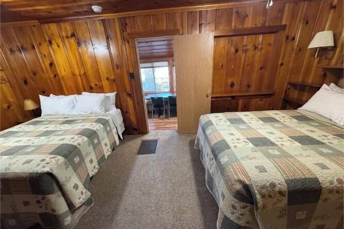 A room with wooden paneling, two beds with quilts, a door leading to another room, and a small table with chairs.