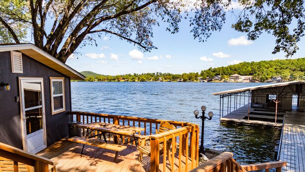 En träveranda med ett bord och stolar, med utsikt över en sjö med en brygga och en byggnad i bakgrunden.