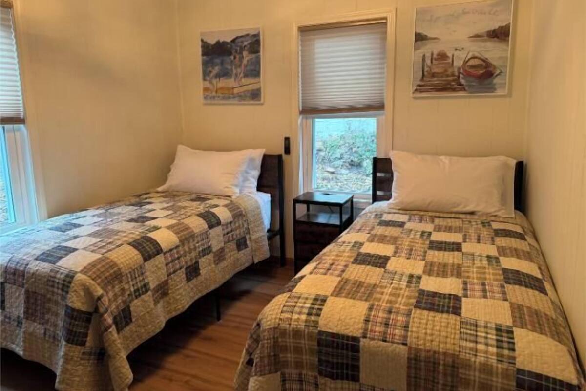 A room with two beds, each with a plaid quilt, a small table, and two paintings on the wall.