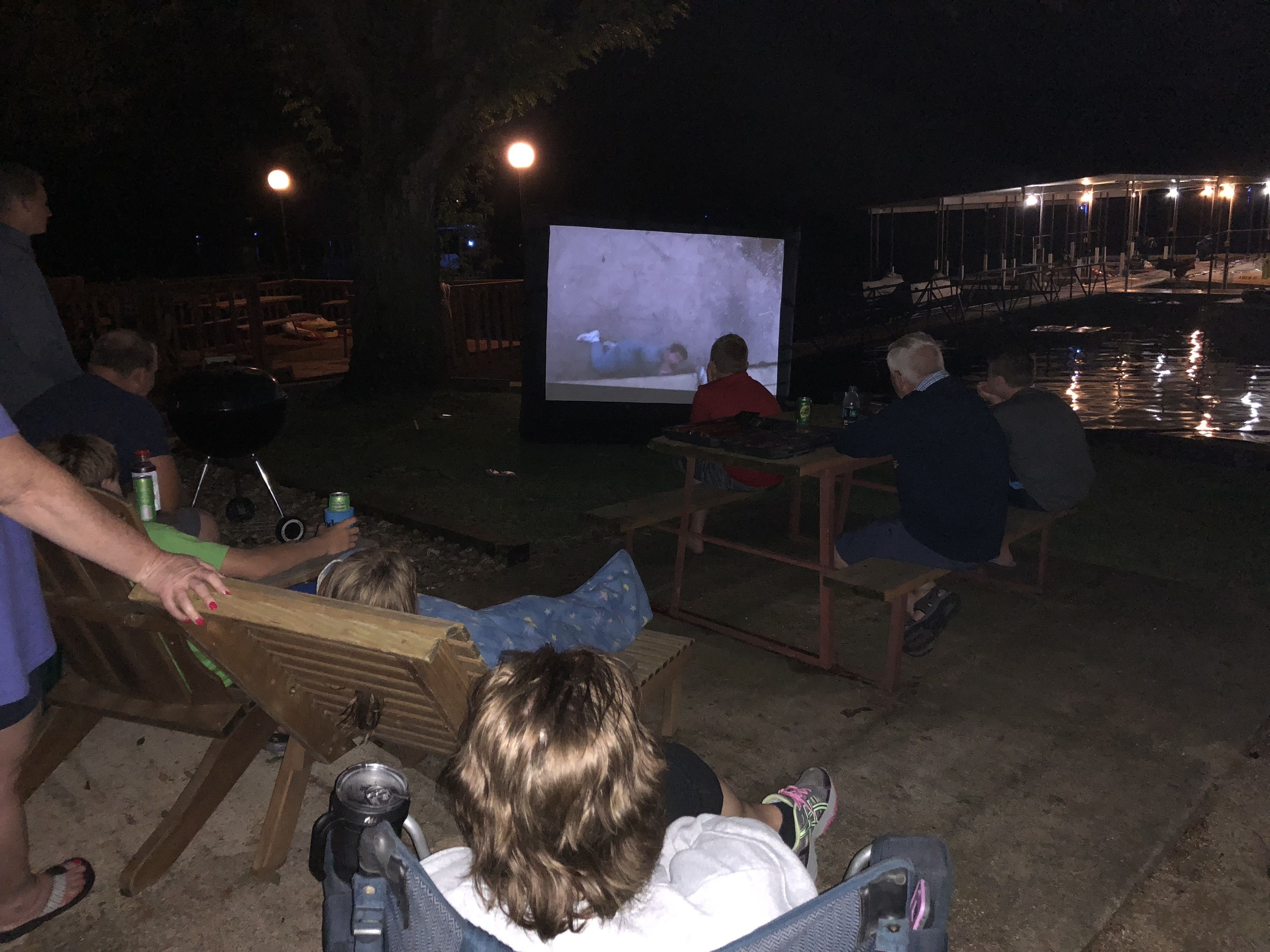 An outdoor gathering with people seated on benches, a large screen displaying a movie, and a body of water in the background.