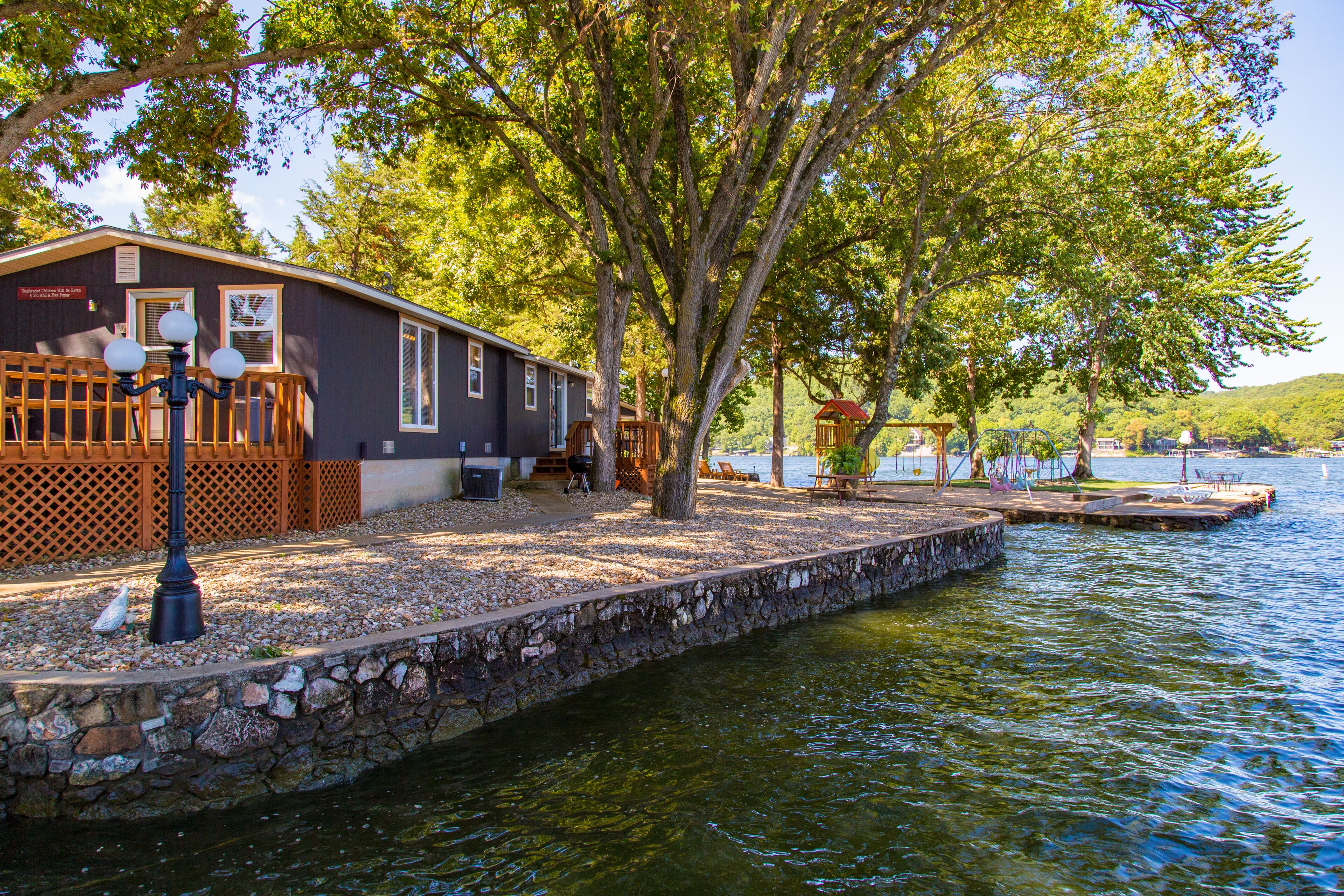 Un chalet au bord du lac, doté d’un quai en pierre qui s’avance dans l’eau, entouré d’arbres et agrémenté d’une terrasse en bois.