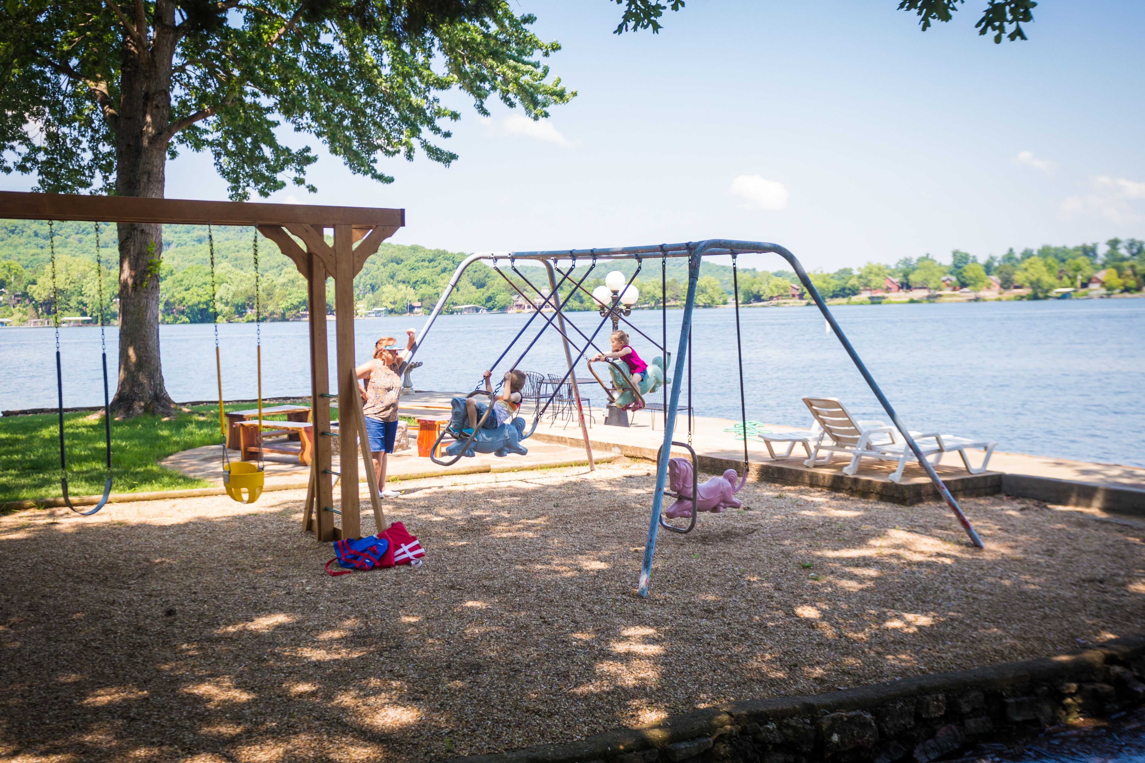 A playground with swings and a swing set, located near a body of water with a tree and benches.