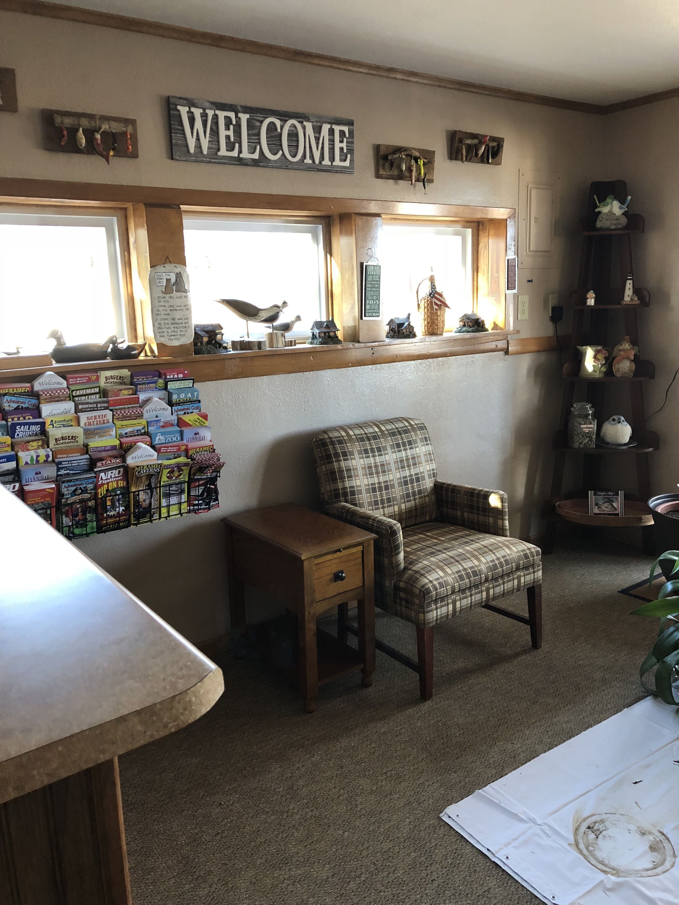 Une pièce chaleureuse avec un fauteuil à carreaux, une petite table d’appoint en bois et une étagère ornée de divers objets décoratifs. Un panneau « BIENVENUE » est accroché au-dessus d’une fenêtre, et un panneau d’affichage avec des brochures de voyage est visible.