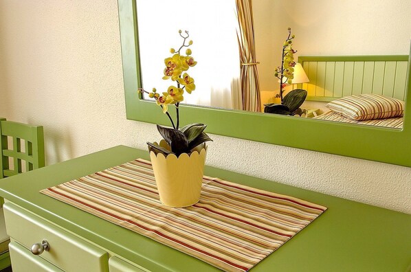 Una habitación con una mesa verde, un mantel a rayas, una planta en maceta, una silla y un espejo que refleja una cama y una lámpara.