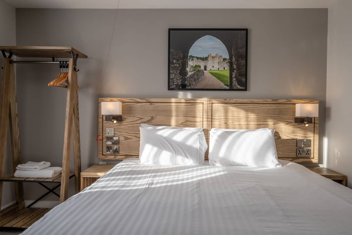 A hotel room with a bed, two bedside lamps, a wooden headboard, a framed picture on the wall, a wooden bench, and a towel rack.