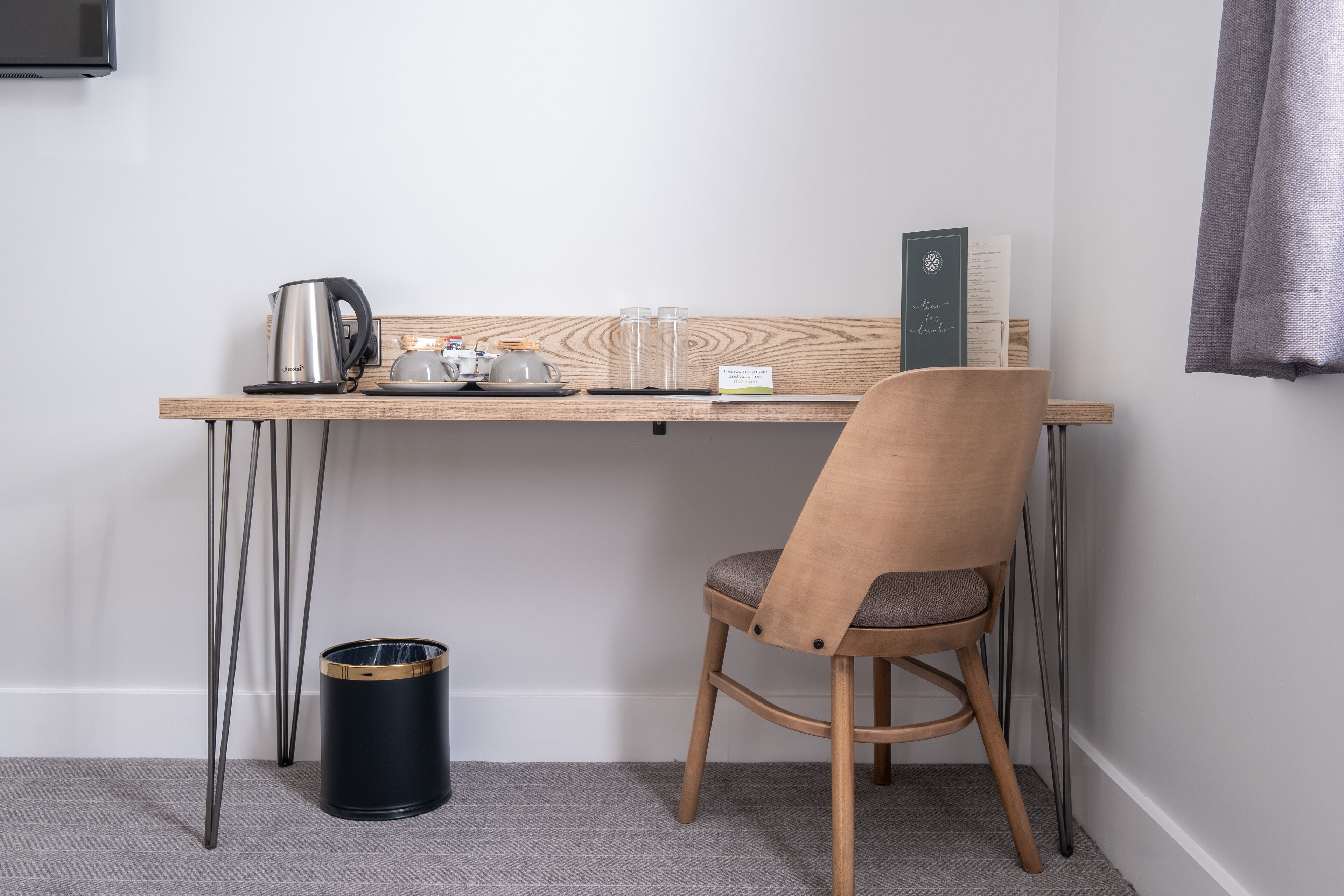 Un petit espace de travail bien aménagé, avec un bureau en bois, une chaise, une bouilloire, une tasse et une poubelle.