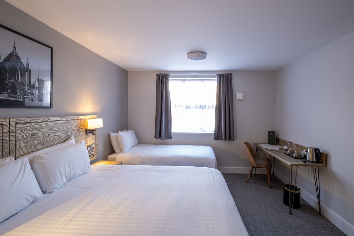 A hotel room with two beds, a desk with a chair, a window with curtains, a wall-mounted lamp, and a framed picture on the wall.